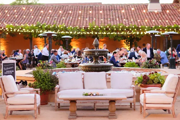 Outdoor patio with white sofas and a lit fountain, guests dining under string lights at dusk