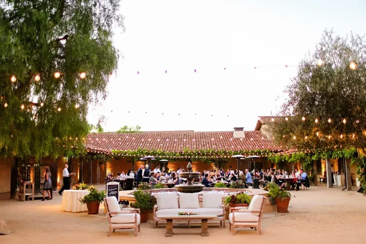Outdoor patio with string lights, white sofas, potted plants, and a tiled-roof restaurant at dusk