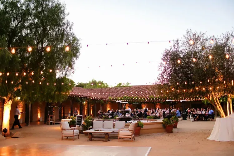 Outdoor courtyard with string lights, trees, and lounge seating at dusk, with people gathered in the background