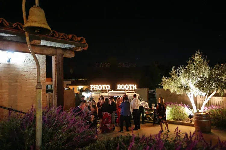 Nighttime crowd gathered at a photo booth beside a lit brick building and tree-lit patio