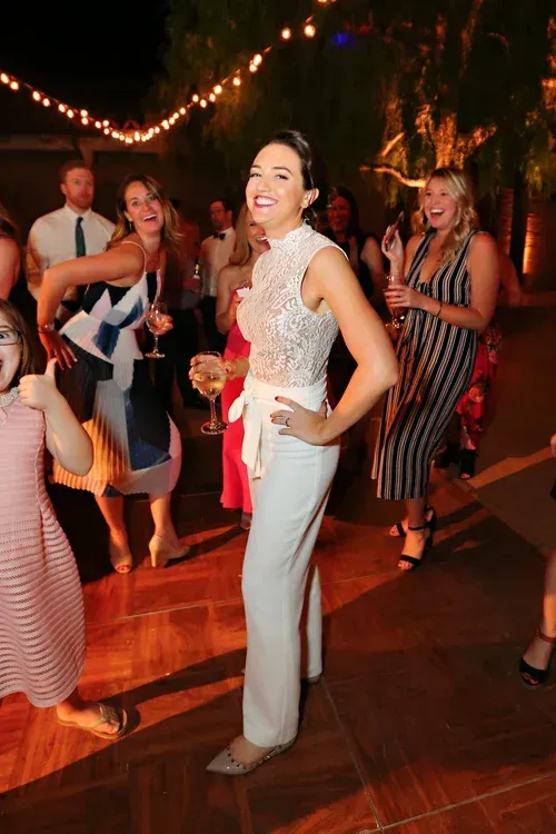 Woman dancing at a party, smiling in a white lace top and light blue pants under string lights.