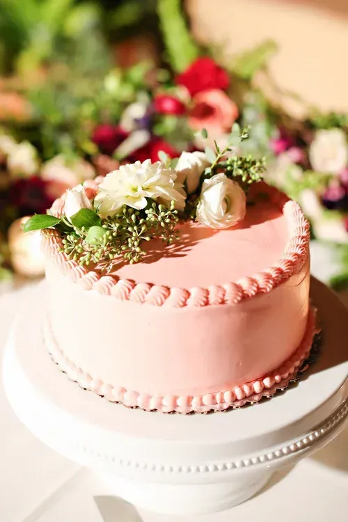 Pink frosted cake with white flowers and greenery on a white stand, in a floral setting