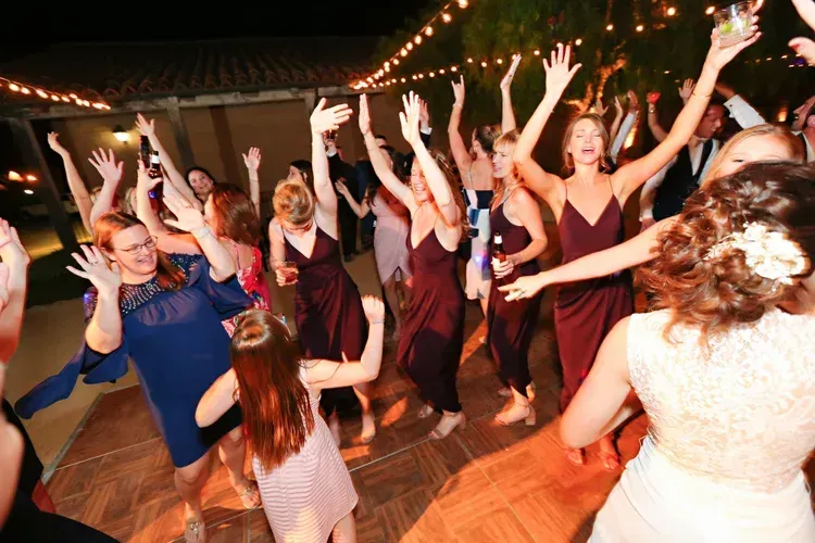 Guests dancing at a lively indoor party under string lights, with arms raised on a wooden floor.