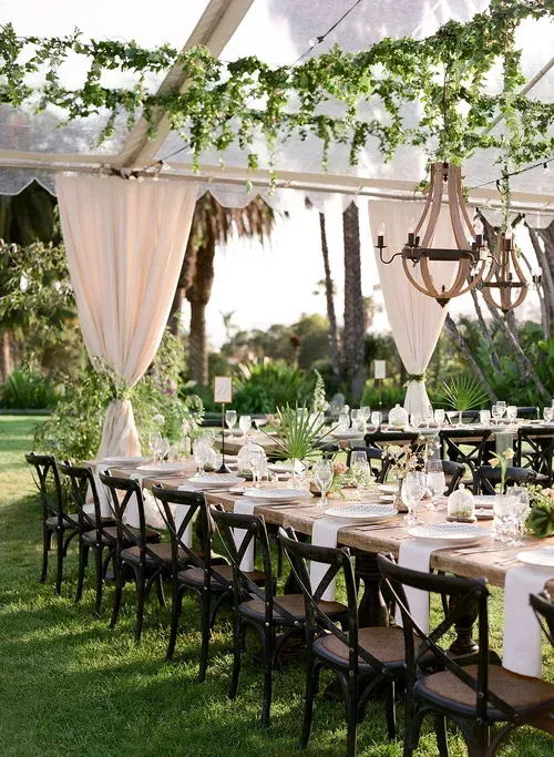 Outdoor wedding reception with long tables, black chairs, white drapes, and greenery under a tent