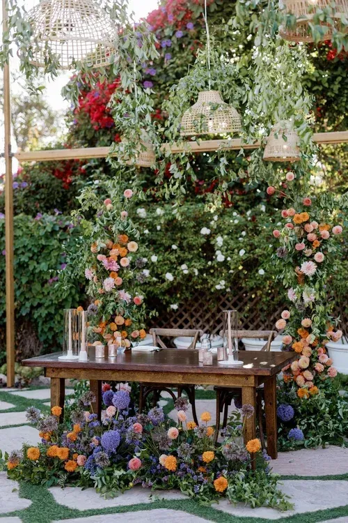 Flower-covered outdoor dining table with colorful floral arch and hanging greenery in a garden setting