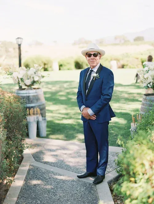 Man in a blue suit and hat standing on a garden path at an outdoor event.
