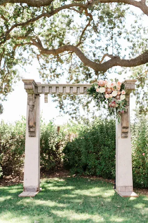 White garden arbor with pink flowers beneath a leafy tree in a sunlit yard