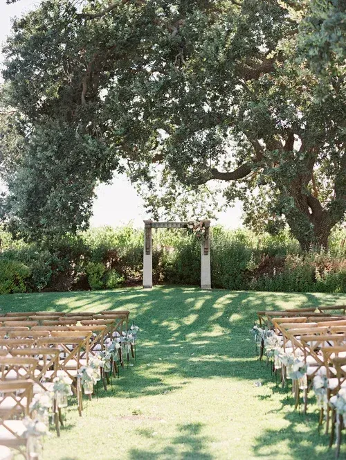 Outdoor wedding ceremony aisle beneath large trees, with rows of chairs and a wooden archway.