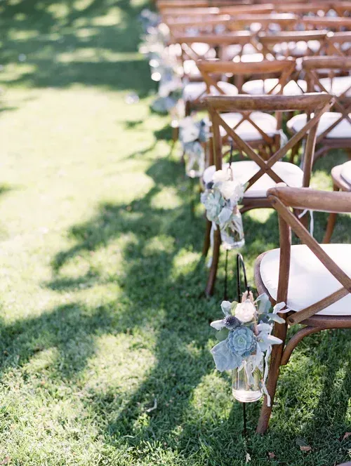 Rows of wooden chairs with white cushions on grass, decorated with white flowers, at an outdoor event