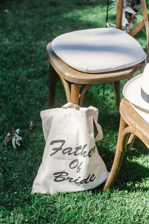 Canvas tote on grass beside white-cushioned chairs, labeled “Father of Bride” at an outdoor event