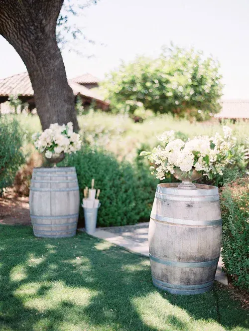 Two wooden barrels with white flowers in a sunny garden beside a tree and lawn