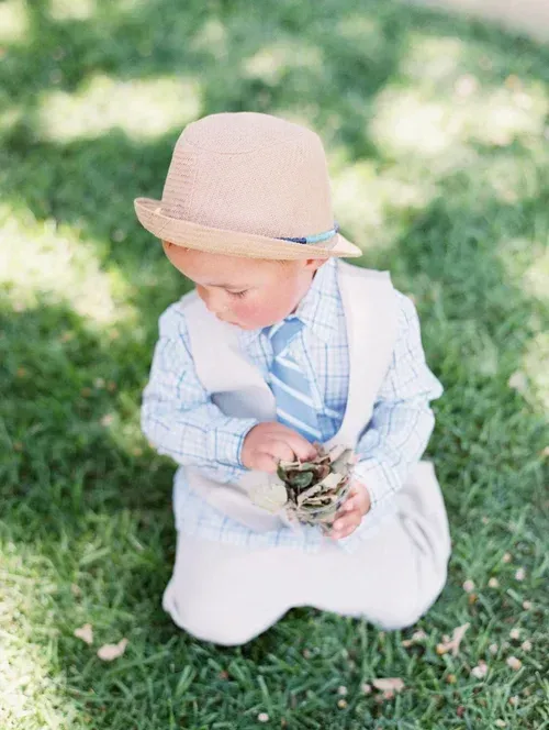 Toddler in a beige hat and light outfit kneeling on grass, holding a small object outdoors.