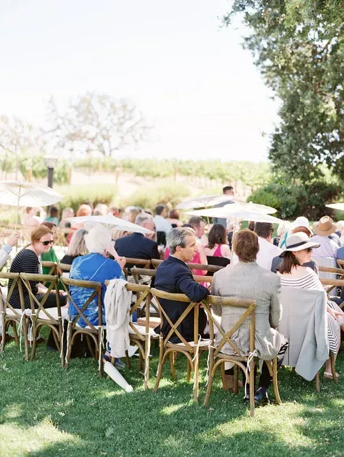 People seated at outdoor tables on a sunny patio with grass, trees, and umbrellas nearby