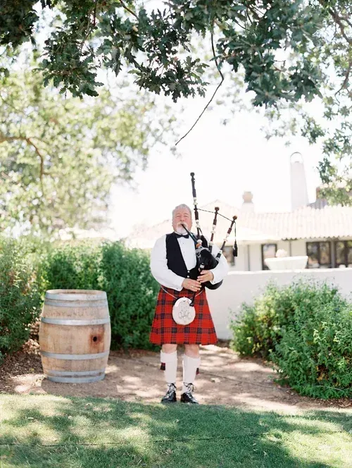 Bagpiper in a red tartan kilt playing outdoors beside a wooden barrel under trees