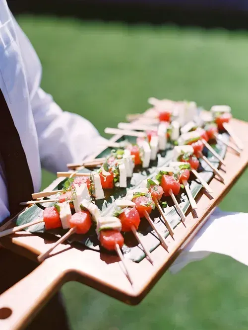 Skewered appetizers with tomatoes and herbs on a wooden serving board, held outdoors on grass