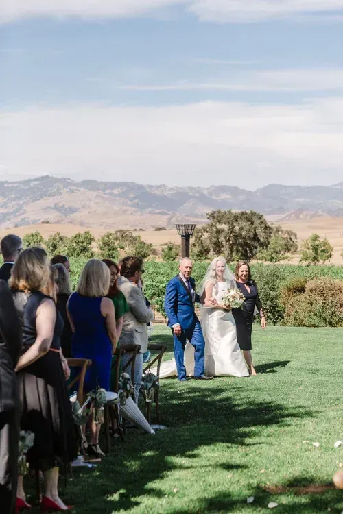 Outdoor wedding guests stand on a lawn with mountains in the background as a bride and groom walk by.