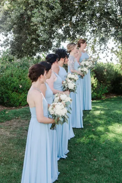 Four women in light blue dresses holding bouquets stand outdoors under a tree at a wedding.