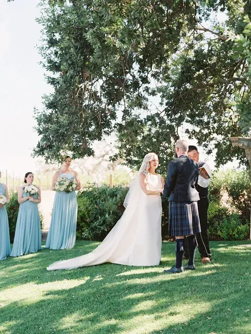 Bride and groom exchanging vows outdoors under a large tree, with bridesmaids nearby