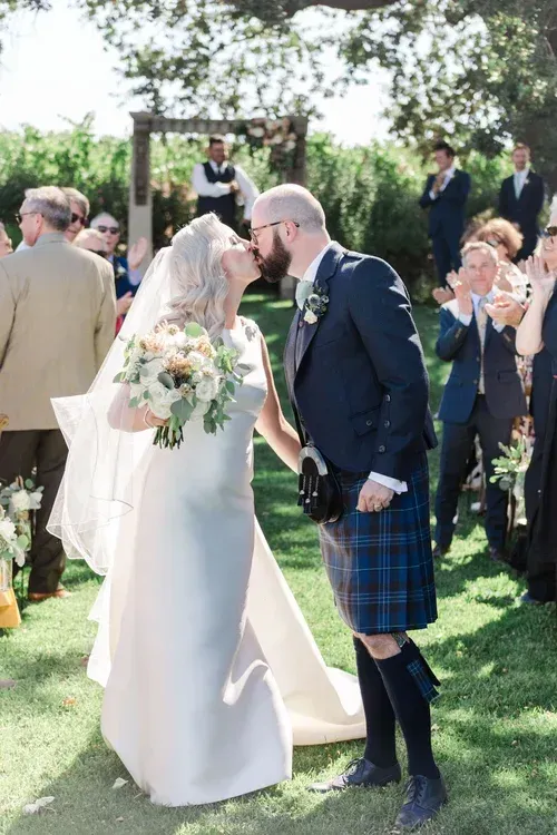 Bride in white dress and groom in navy kilt kiss outdoors at a wedding ceremony