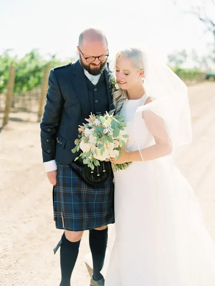 Bride and groom in wedding attire walking outdoors, holding a bouquet in a bright vineyard setting