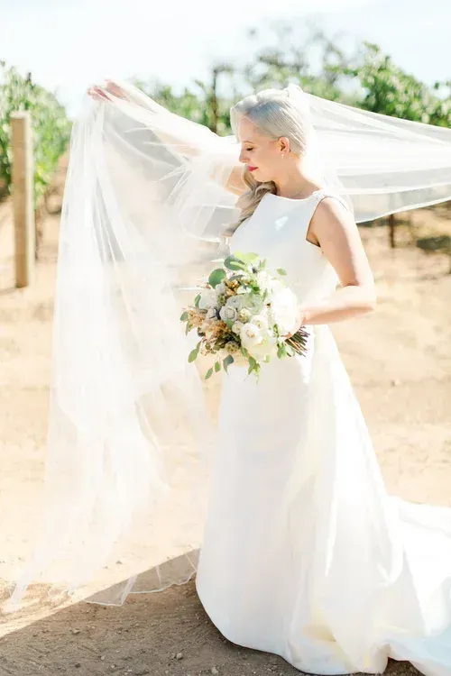 Bride in a white dress holding a bouquet with her veil blowing in the wind outdoors