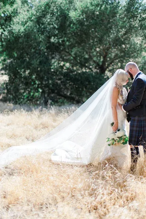 Bride and groom stand together in a sunny field, her long veil trailing in the grass.