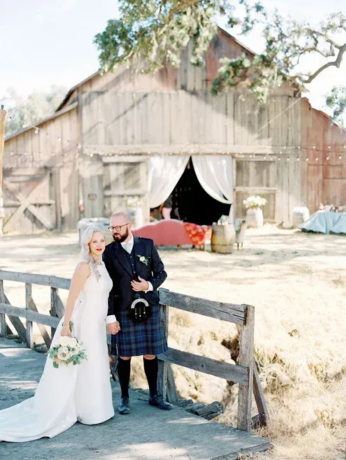 Bride and groom posing in front of a rustic barn with white drapes and a red car inside