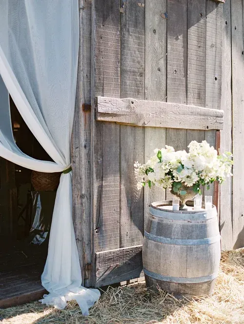 Rustic wooden barrel with white flowers beside a barn door and draped curtain