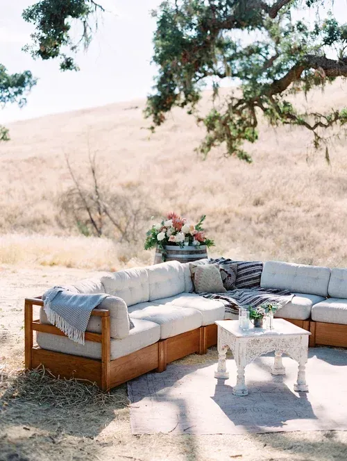 Outdoor patio seating with white cushions, wooden sofa, small table, and flowers under a tree in a sunny field