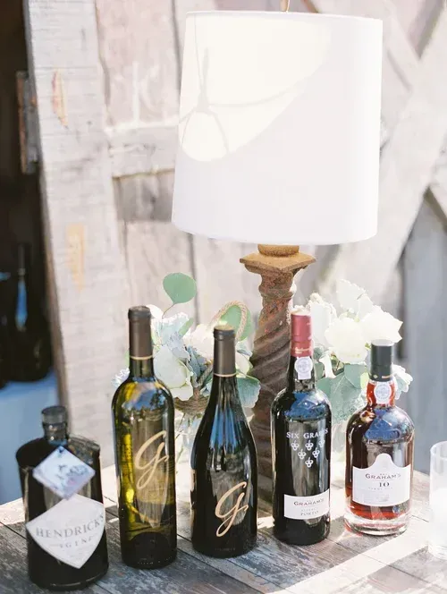 Assorted wine bottles arranged on a table with a white lamp and decorative flowers in the background