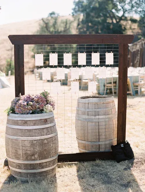 Rustic outdoor wedding photo backdrop with wooden frame, string lights, and flower-filled barrel table
