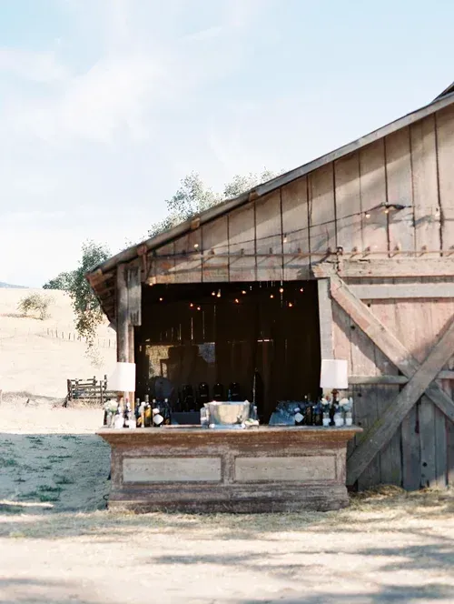 Rustic wooden barn with open stall, bottles on a counter, and sandy ground outside