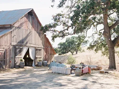 Rustic barn with open doorway, fenced pens, and a large tree in a sunny rural yard.