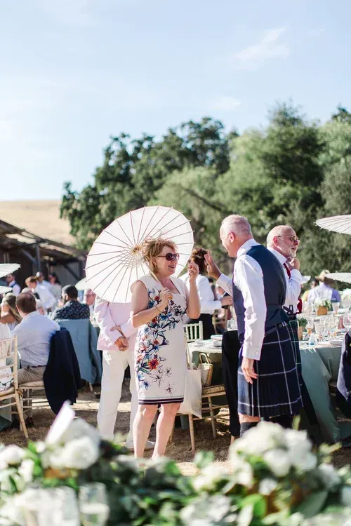 Outdoor wedding reception with guests in formal attire, including a woman holding a parasol and a man in a kilt.