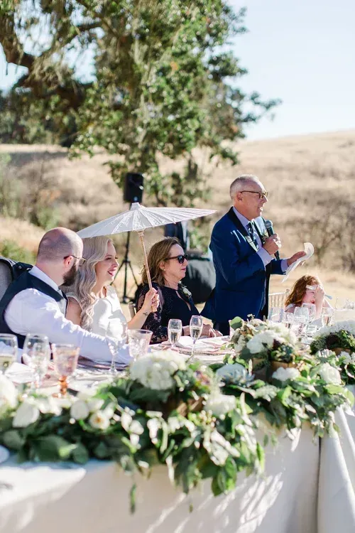 Outdoor wedding reception with a man speaking at a decorated table under an umbrella, guests seated nearby
