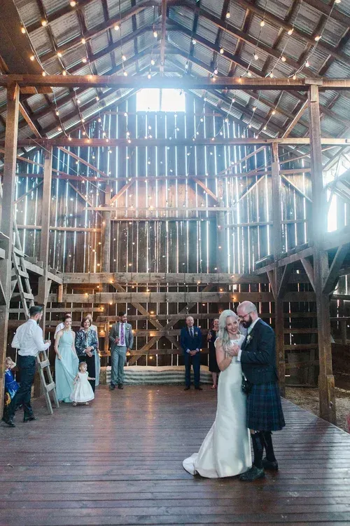 Couple dancing in a rustic barn venue beneath a high wooden roof, with guests nearby