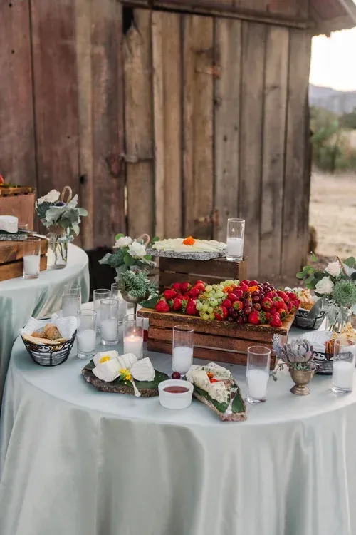 Rustic outdoor dessert table with cakes, berries, flowers, and glassware against a wooden barn backdrop