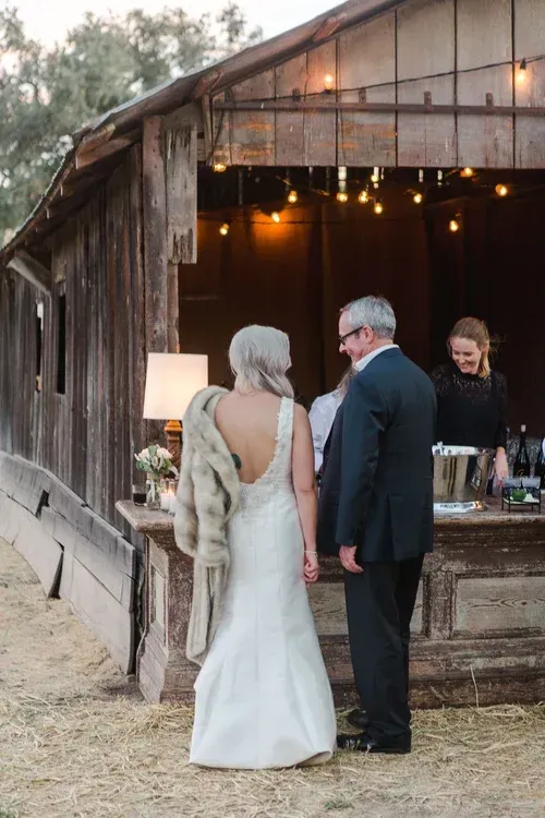 Wedding couple standing at a rustic barn reception entrance, facing a bar under warm string lights
