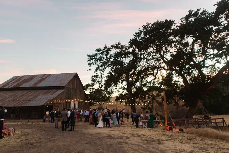 People gathered outside a rustic barn at dusk under a tree, with warm lights and a dirt yard.