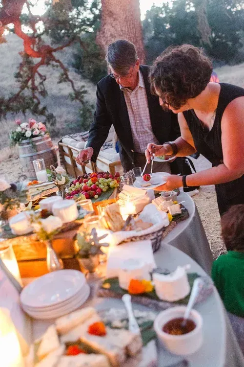People serving food at a candlelit outdoor dinner table with dishes, drinks, and flowers