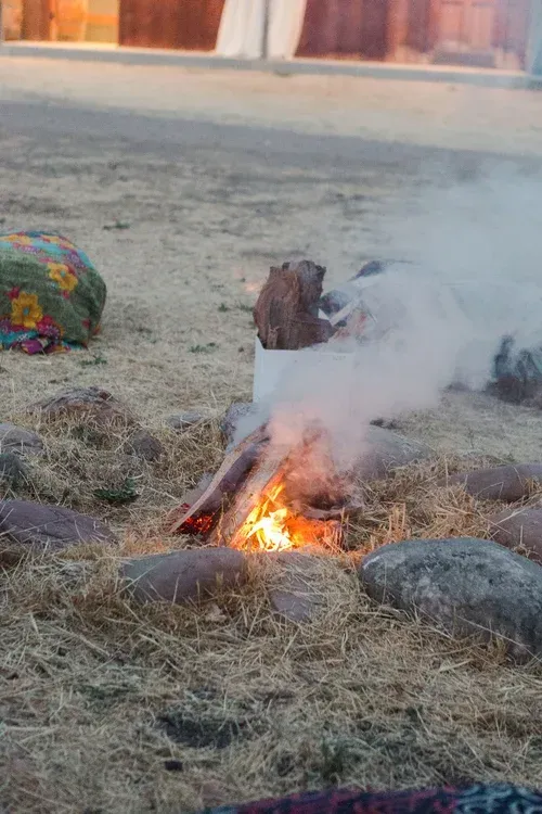 Small campfire burning in dry grass with smoke rising near rocks and a building in the background