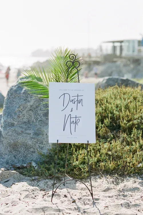 White sign on a stand in beach grass reading “Dusty Hite” beside a green palm frond.