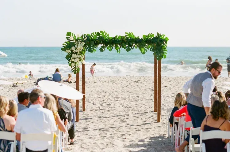 Beach wedding ceremony with guests seated under a leafy arch by the ocean