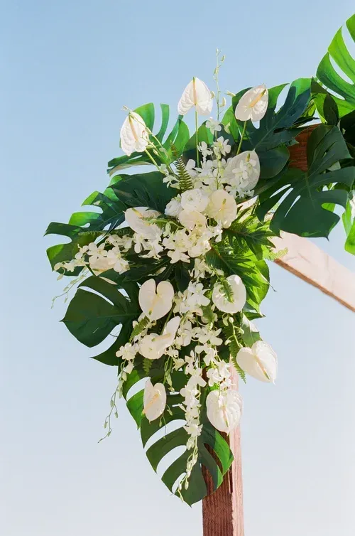 White and green floral arch arrangement with monstera leaves against a blue sky