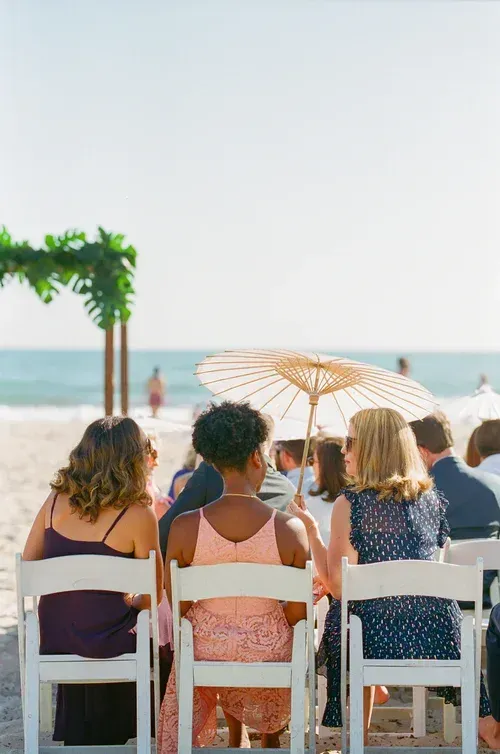 People seated on beach chairs under an umbrella, facing the ocean on a sunny day