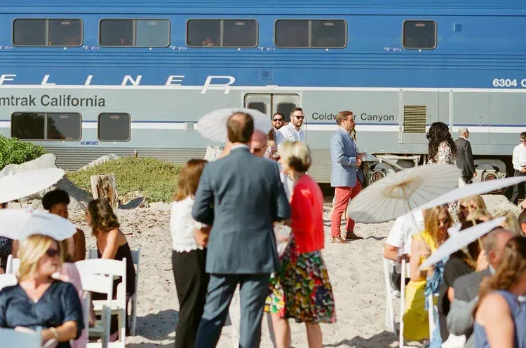 People gathered on a platform beside a blue train at a station, with mountains in the background.