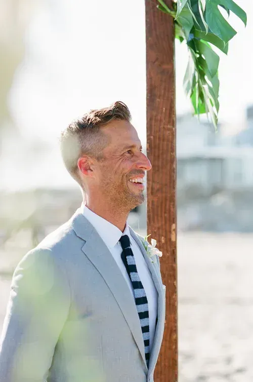 Smiling man in light blazer and striped shirt standing outdoors near a wooden post and greenery