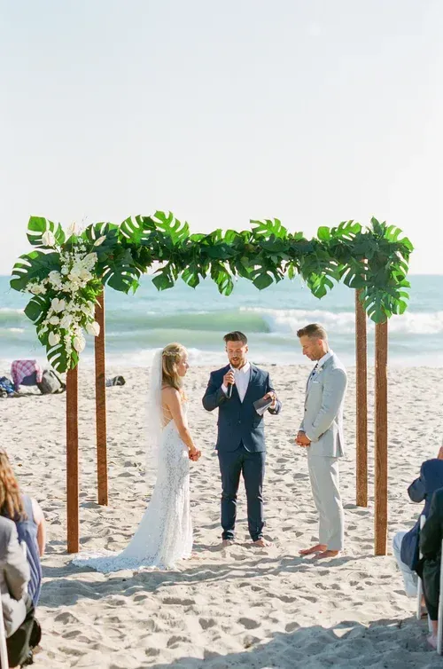 Beach wedding ceremony under a leafy arch, with officiant and couple standing on the sand by the ocean.