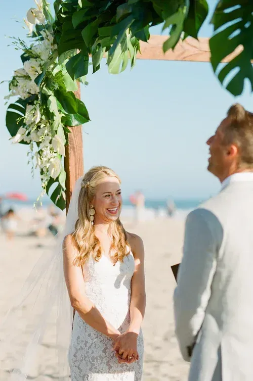 Bride smiling under leafy wedding arch on a sunny beach, facing groom