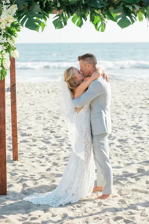 Couple embracing on a sunny beach beneath green foliage, with ocean waves in the background